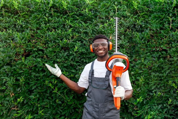 Trabalhador de jardim em uniforme corta arbustos homem afro-americano em fones de ouvido segura escova elétrica