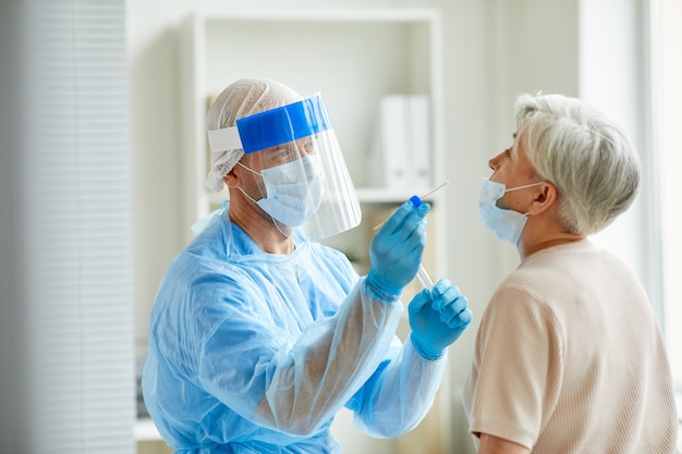 Trabalhador de laboratório moderno usando equipamento de proteção individual testando mulher idosa para coronavírus usando método de cotonete nasal