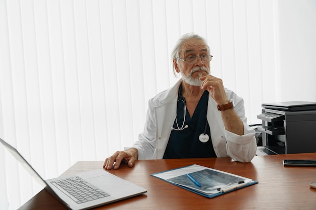 Trabalhador de saúde sênior fazendo alguma papelada e usando laptop enquanto trabalhava no consultório médico