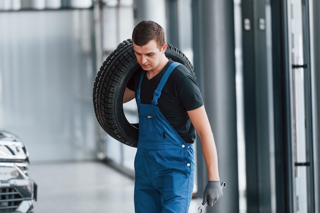 Trabalhador de uniforme preto e azul segurando a roda do carro e tem trabalho dentro de casa