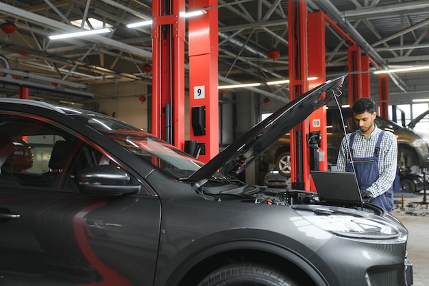 Trabalhador do gerente mecânico do homem mecânico usando um computador portátil verificando o carro na oficina no centro de serviços de reparação de automóveis Jovem engenheiro olhando os detalhes do veículo de inspeção sob o capô do carro