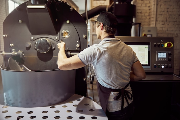 Trabalhador do sexo masculino com avental operando equipamento profissional para torrar grãos de café enquanto está de pé junto ao painel de controle