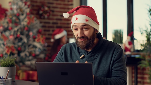 Trabalhador do sexo masculino falando em conferência de videochamada com webcam, usando chapéu de Papai Noel durante a véspera de Natal. Homem conversando na chamada de videoconferência on-line no escritório com decorações de Natal.