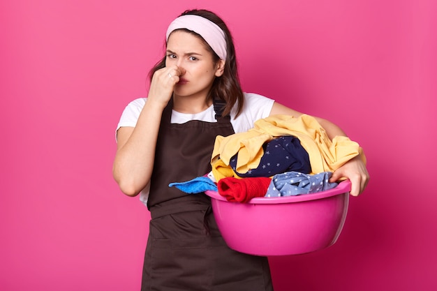 Trabalhador doméstico morena segurando o nariz com uma mão, tendo bacia rosa cheia de roupas com cheiro ruim, tentando suportar o cheiro. poses de modelo exausto emocional isoladas sobre parede rosa.