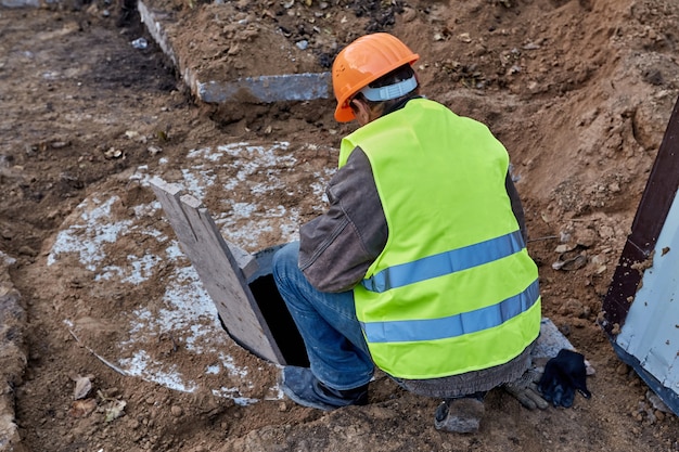 Trabalhador está olhando para uma escotilha de esgoto para verificar as comunicações que conduzem a um novo edifício alto em construção close-up vista traseira