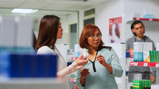 Trabalhador farmacêutico mostrando frasco de comprimidos de cardiologia para cliente asiático em farmácia, explicando o tratamento para curar problemas cardíacos. Farmacêutica falando com mulher sobre produtos médicos.