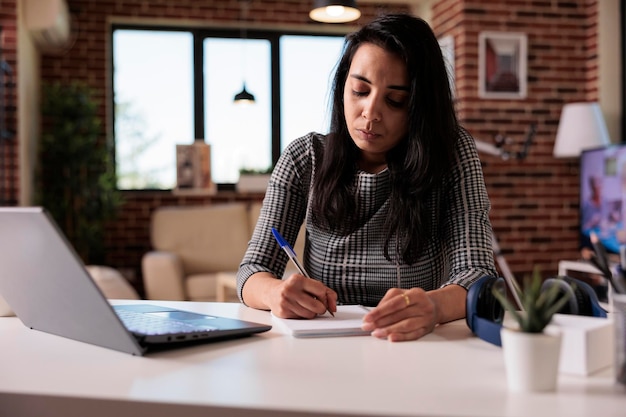 Trabalhador indiano tomando notas de papelada no caderno, escrevendo relatório de informações ou lição de classe escolar. Trabalhando no laptop com o livro didático para fazer pesquisas on-line e planejar a apresentação na web.