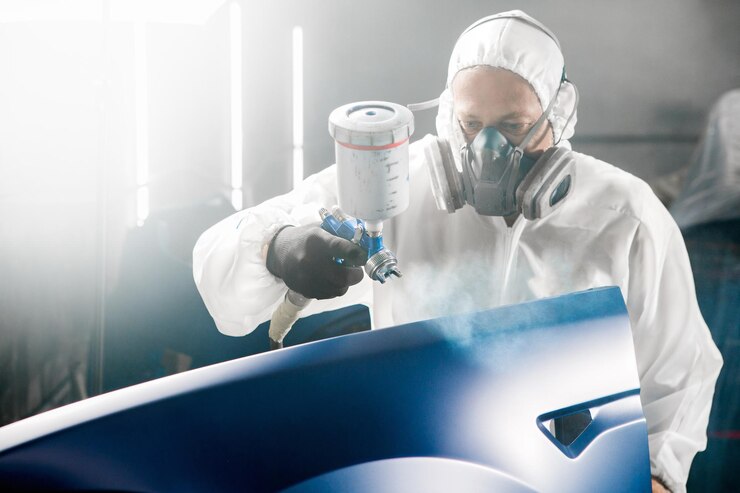 Foto trabalhador industrial masculino pulveriza tinta usando uma pistola de aerógrafo em parte do carro usando uma máscara protetora