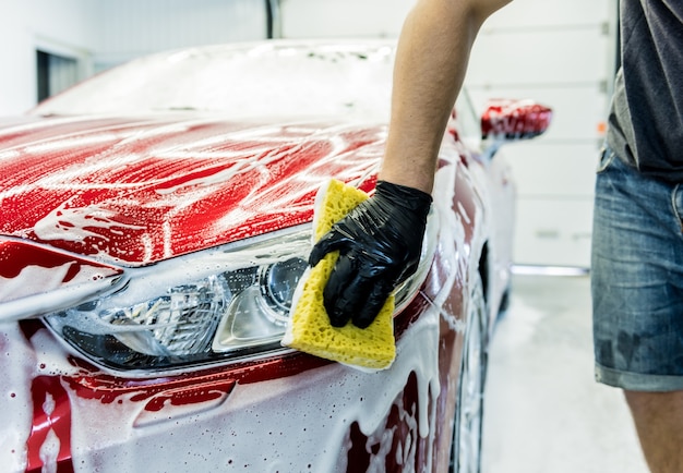 Trabalhador lavando carro vermelho com esponja em uma lavagem de carro.