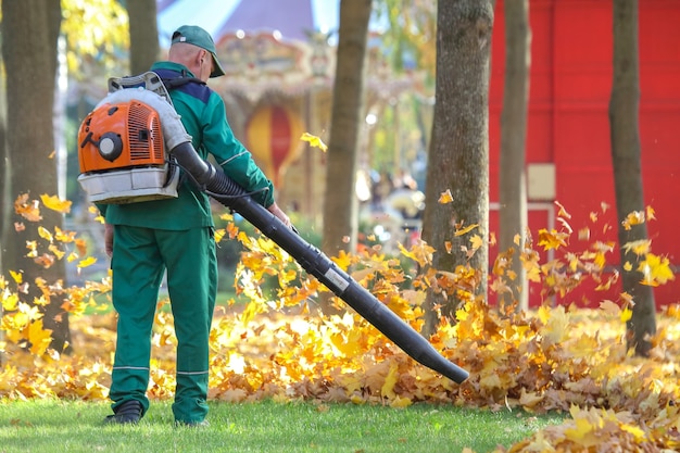 Trabalhador limpa folhas de outono com um ventoinha em um parque da cidade