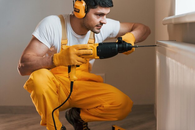 Trabalhador manual de uniforme amarelo trabalha dentro de casa usando broca de martelo Concepção de renovação de casa