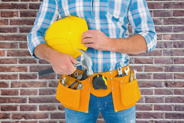 Trabalhador manual usando cinto de ferramentas enquanto segura o martelo e o capacete contra a parede de tijolos vermelhos