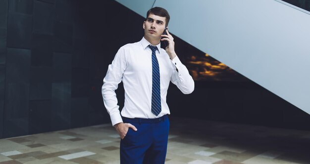 Trabalhador masculino adulto pensativo em elegante camisa branca e gravata azul em pé tendo telefonema no salão de mármore do prédio comercial olhando para longe