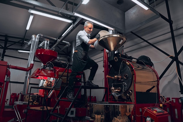 Trabalhador masculino bonito no avental despejando grãos de café verde no funil de metal Vista inferior