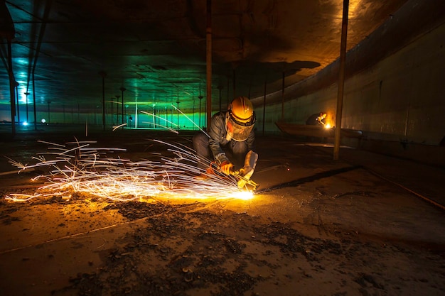 Trabalhador masculino corte de metal faísca na placa de aço do fundo do tanque com flash de luz de corte de perto, use luvas de proteção e máscara em espaço confinado lateral.