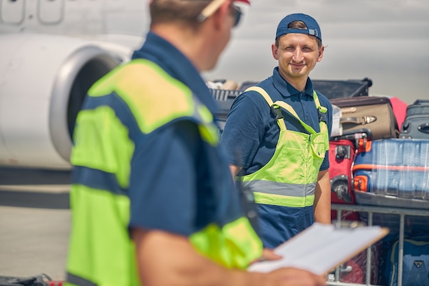 Trabalhador masculino de uniforme sorridente, olhando para seu colega com uma prancheta na mão