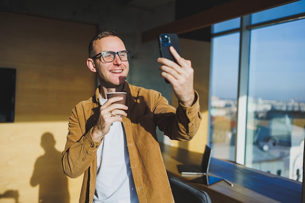 Trabalhador masculino sorridente em roupas casuais, olhando para longe enquanto fala no celular e bebe café durante o intervalo no espaço de trabalho criativo moderno
