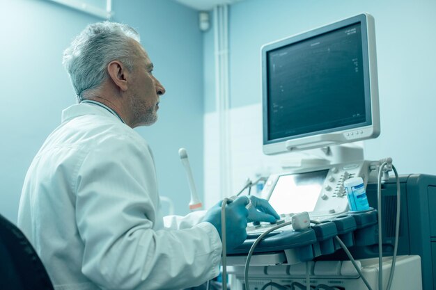 Trabalhador médico maduro em luvas de borracha, tocando os botões de um equipamento de ultrassom e olhando para a tela