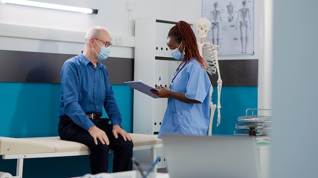 Trabalhador médico tomando notas na consulta com o paciente antigo, usando máscaras faciais no escritório. Enfermeira feminina consultando homem doente na consulta de check-up, clínica de apoio à saúde.