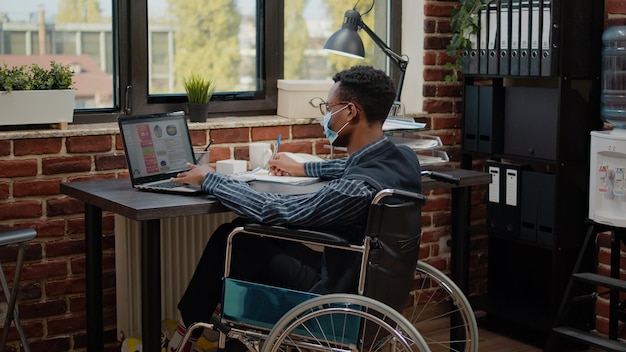 Trabalhador sentado em cadeira de rodas e planejamento de projeto de negócios para crescimento financeiro, usando informações comerciais no laptop. Funcionário da empresa com deficiência crônica trabalhando e usando máscara facial.