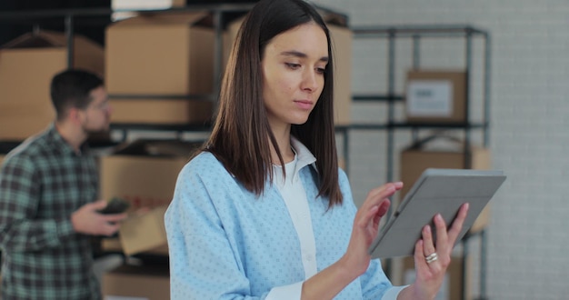 Trabalhador verificando notas de escrita de inventário no tablet Uma mulher trabalha em um armazém com fileiras de prateleiras com pacotes de caixas