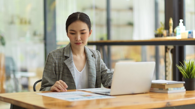 Trabalhadora de escritório asiática focada, sentada à mesa, trabalhando em um projeto, digitando no laptop, concentrada em relatórios financeiros, trabalhando em um espaço de co-working em um café