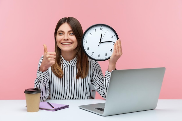 Trabalhadora de escritório de mulher feliz positiva mostrando os polegares para cima segurando o relógio de parede grande, olhando para a câmera com um sorriso, gerenciamento de tempo. Tiro de estúdio interior isolado no fundo rosa