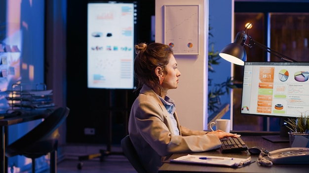 Trabalhadora de escritório feminina verificando estatísticas de negócios à noite digitando plano de pesquisa no pc na mesa. Empreendedor analisando diagramas e relatórios financeiros, trabalhando no progresso e desenvolvimento.