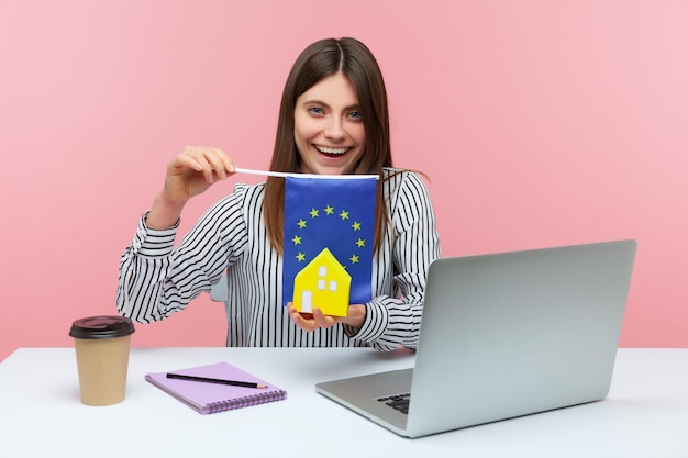 Trabalhadora de escritório sorridente positiva segurando modelo de casa de papel e bandeira da união europeia sonhando em viver no país europeu sentado no local de trabalho Estúdio interior isolado no fundo rosa