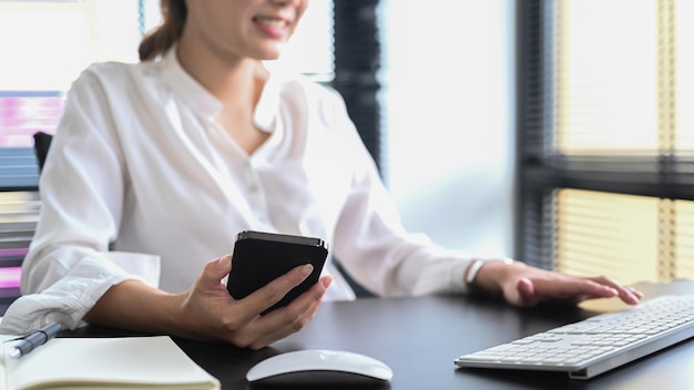 Trabalhadora de escritório sorridente segurando telefone inteligente e digitando no teclado sem fio Tiro recortado