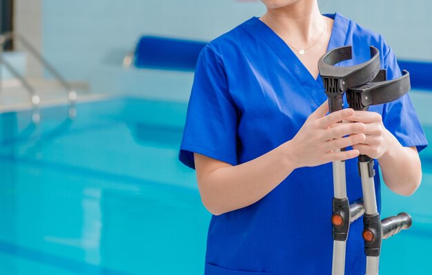 Trabalhadora hospitalar caucasiana com muletas de reabilitação nas mãos piscina no fundo