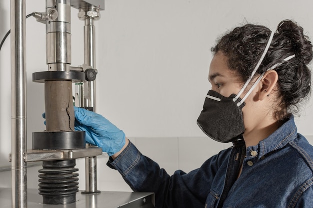 Trabalhadora industrial com máscara protetora e luvas de látex azuis testando um cilindro de argila em laboratório geológico