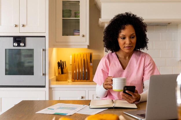 Trabalhadora médica biracial usando smartphone e laptop, trabalhando na cozinha, espaço de cópia. Serviços médicos, trabalhando em casa, trabalho remoto, comunicação, estilo de vida e vida doméstica, inalterados.