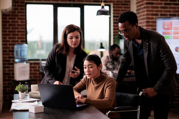 Trabalhadores da empresa fazendo trabalho em equipe no escritório amigável para deficientes, usando laptop para navegar na internet. Funcionária usuária de cadeira de rodas com deficiência física trabalhando na estratégia do projeto.