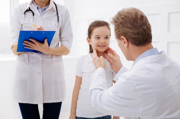 Trabalhando com crianças. pediatra profissional gentil e qualificado examinando o pescoço da menina enquanto seu assistente fica por perto e observa o que ele diz