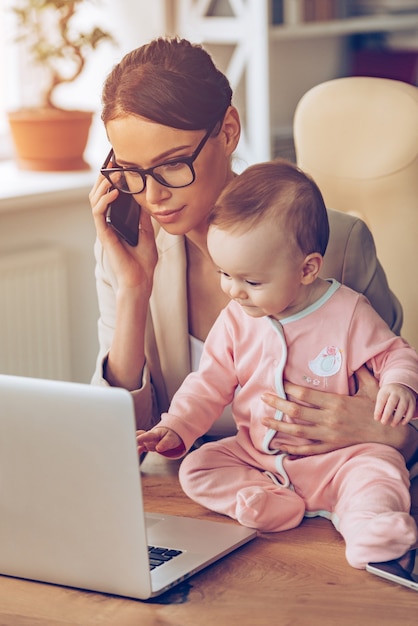 Trabalhando com o bebê. bela jovem empresária falando no celular e olhando para o laptop enquanto está sentada com sua filha no local de trabalho