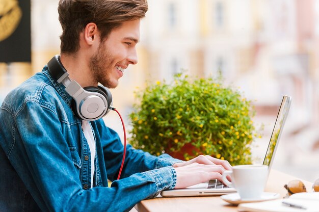 Trabalhando com prazer. Vista lateral de um jovem sorridente trabalhando em um laptop