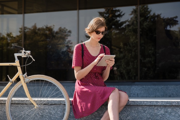 Trabalhando com tecnologia fora do escritório. mulher jovem em um elegante vestido retrô, trabalhando com um computador tablet ao ar livre em uma área urbana, sentada ao lado de uma bicicleta.