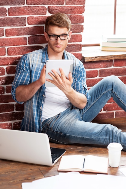 Trabalhando em casa. Jovem bonito sentado no chão e trabalhando em um tablet digital enquanto um laptop e documentos estão perto dele