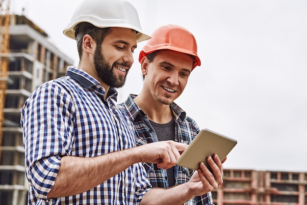Foto trabalhando em equipe, dois jovens e alegres construtores em capacetes de proteção estão usando tablet digital