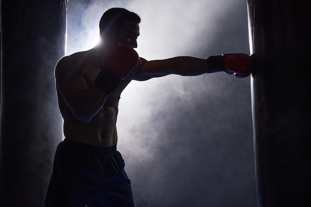 Trabalhando em sua técnica Foto recortada de um jovem boxeador masculino em silhueta malhando em um saco de pancadas no ginásio