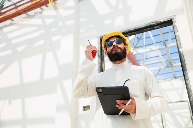 Trabalhando em tablet e walkie talkie Homem árabe Engenheiro civil de construção usa computador tablet digital e usando capacete de segurança trabalhando no canteiro de obras