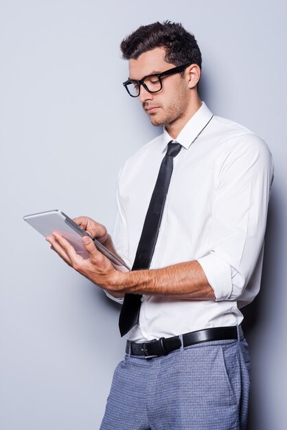 Trabalhando em um tablet digital. Jovem bonito de camisa e gravata trabalhando em um tablet digital em pé contra um fundo cinza