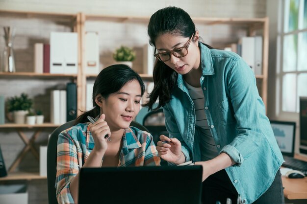 Trabalhando juntos no projeto. Duas jovens mulheres asiáticas confiantes olhando para monitor de laptop e notebook discutindo novo plano de cooperação no escritório do estúdio. meninas elegantes no local de trabalho moderno falando.