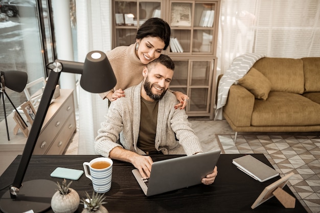 Trabalhando no laptop. Casal sorridente e positivo se abraçando suavemente enquanto olham para a tela do laptop