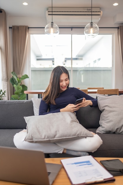 Trabalhe a partir do conceito de casa, uma jovem empresária sentada em um sofá aconchegante, usando seu telefone celular, conversando com os clientes.