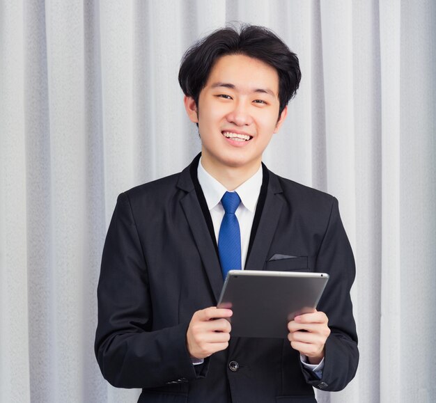Trabalhe em casa, o jovem empresário asiático usa terno, videoconferência ou facetime, ele sorrindo usando um tablet digital inteligente tocando na tela do escritório em casa, olhando para a câmera