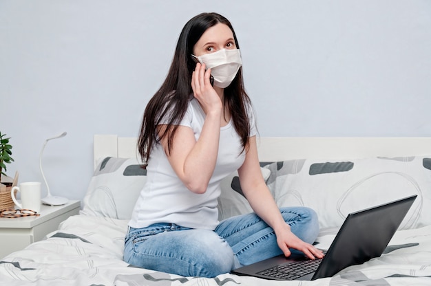 Trabalhe em casa ou a distância, em quarentena. Mulher em máscara protetora trabalhando remotamente na cama usando o laptop e falando no telefone.