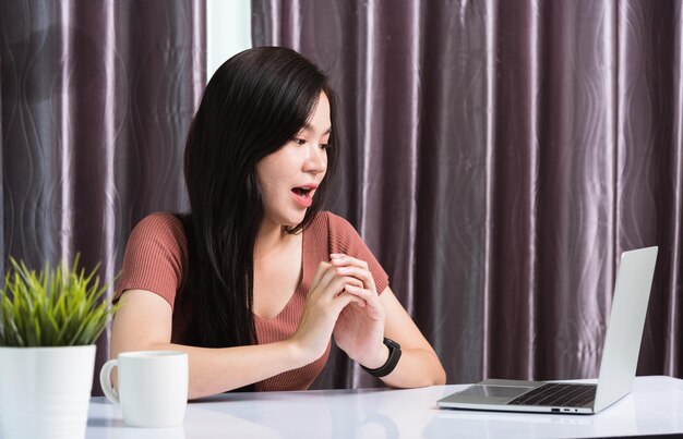 Trabalhe em casa, Sorrindo feliz negócios asiáticos jovem mulher bonita sentada na mesa de trabalho videoconferência por webcam tecnologia de computador portátil em escritório doméstico, expressão facial WOW assistindo