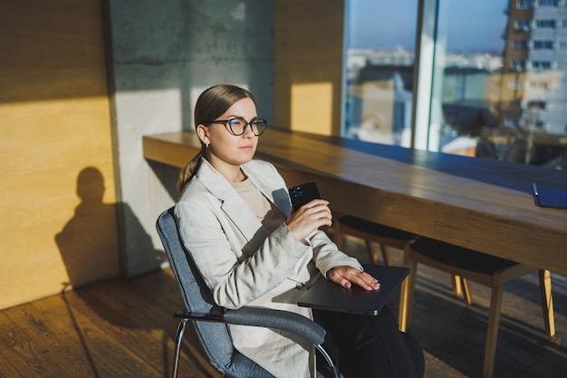 Trabalhe em um escritório moderno Uma jovem empresária positiva em roupas casuais e óculos, sentada no escritório com um telefone celular enquanto trabalha em um projeto remoto usando um laptop